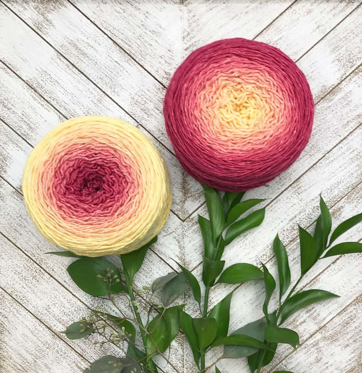 Two gradient yarn cakes in yellow to pink tones with greenery on white wood background Wonderland Yarns Blossoms gradient yarn