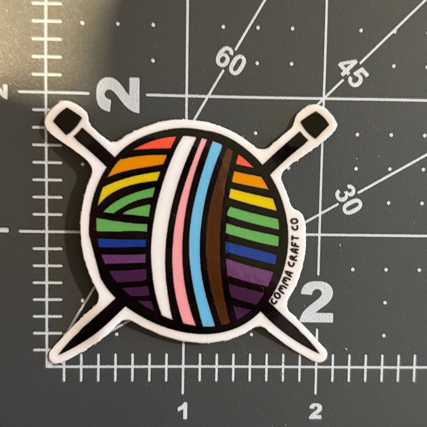 Comma Craft Co intersectional pride knitting sticker featuring colorful striped yarn with knitting needles