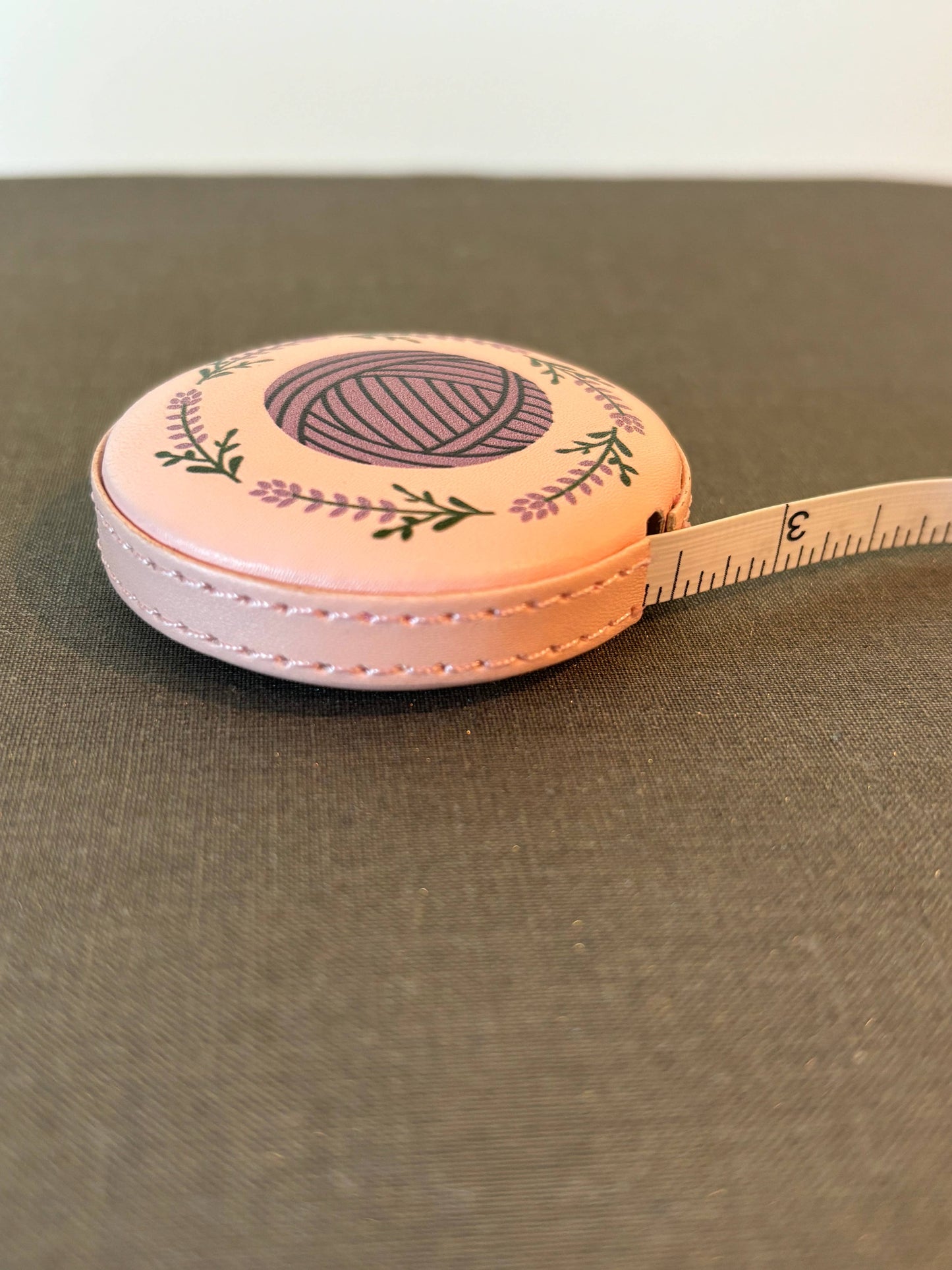 Pink round retractable cute tape measure for crafters with yarn ball and floral design showing inch markings