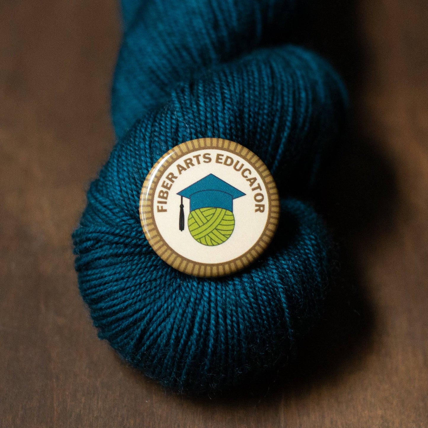 Camp Stitchwood Fiber Arts Educator Button on a teal yarn skein for fiber arts enthusiasts