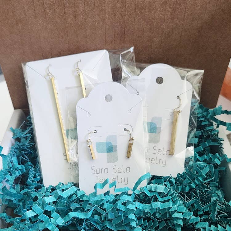 Sara Sela Jewelry packaged gold bar dangle earrings on branded cards with teal crinkle filler crescent rainbow moonstone earrings