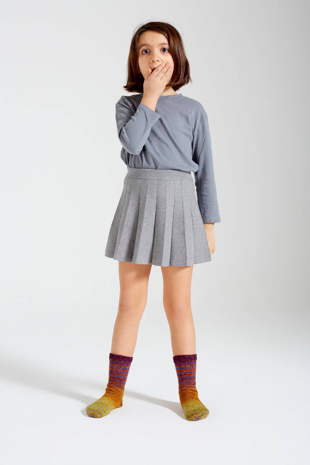Child wearing knitted socks made from Urth Yarns Uneek Sock Kit Mini, styled with a gray top and skirt.