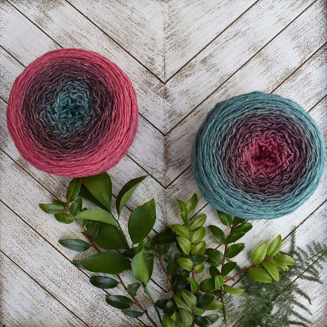 Two hand dyed yarn cakes in teal and rose on whitewashed chevron wood with greenery Wonderland Yarns Blossoms gradient yarn