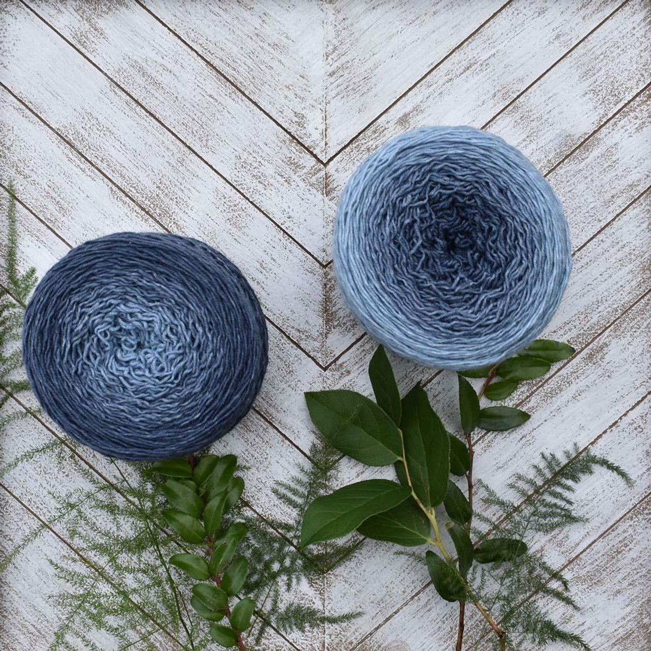 Two blue gradient yarn cakes showcasing Wonderland Yarns Blossoms gradient yarn on white chevron wood with green sprigs