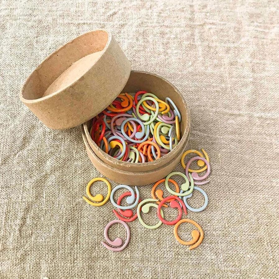 Colorful Cocoknits split ring stitch markers in a round cardboard tin on a natural linen background