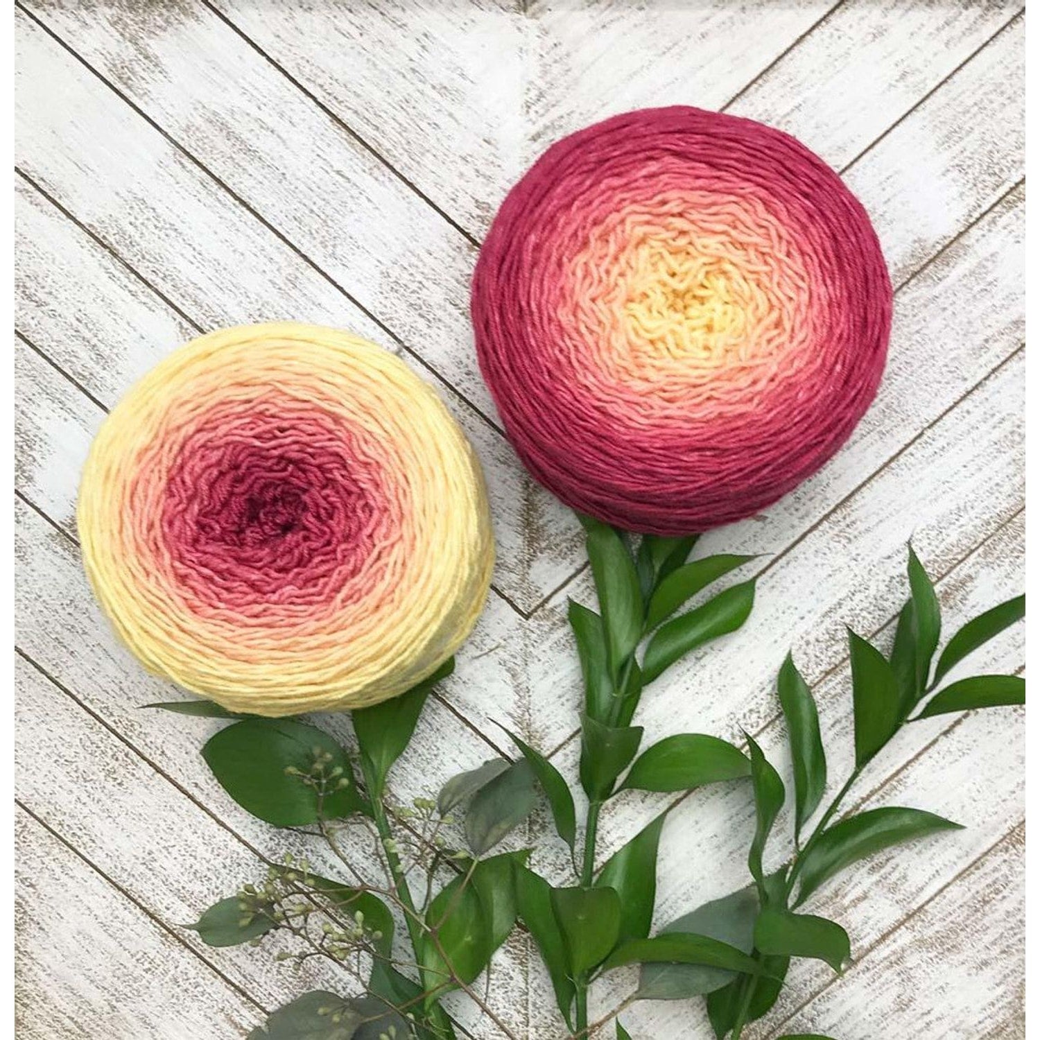 Two gradient yarn cakes in yellow to pink tones with greenery on white wood background Wonderland Yarns Blossoms gradient ya…