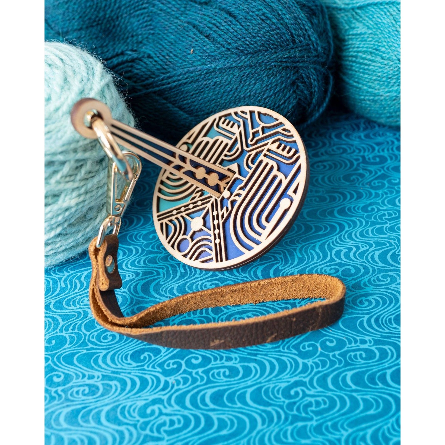 lemonwood art deco stitch marker blue enamel and brass disc with leather strap next to teal and aqua yarn for knitters