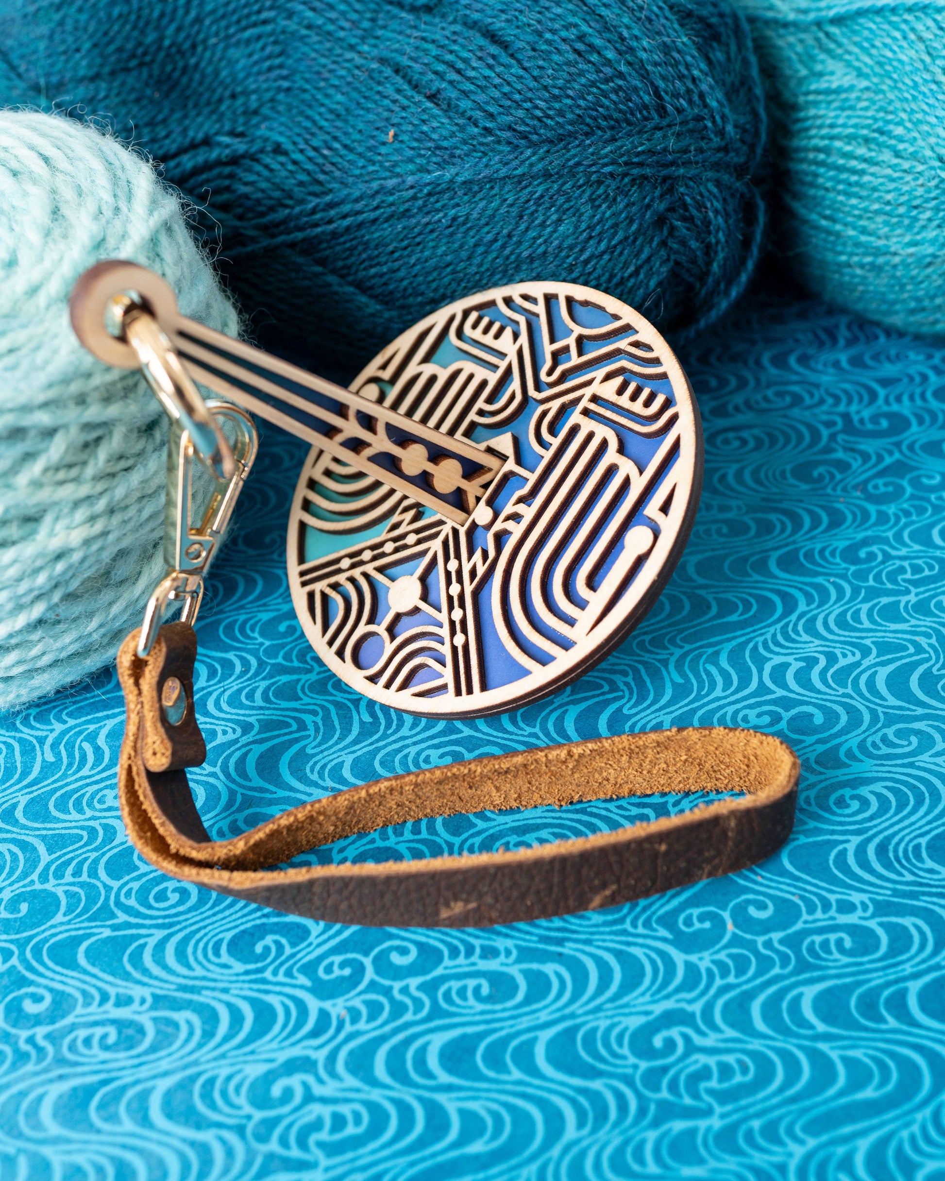 lemonwood art deco stitch marker blue enamel and brass disc with leather strap next to teal and aqua yarn for knitters