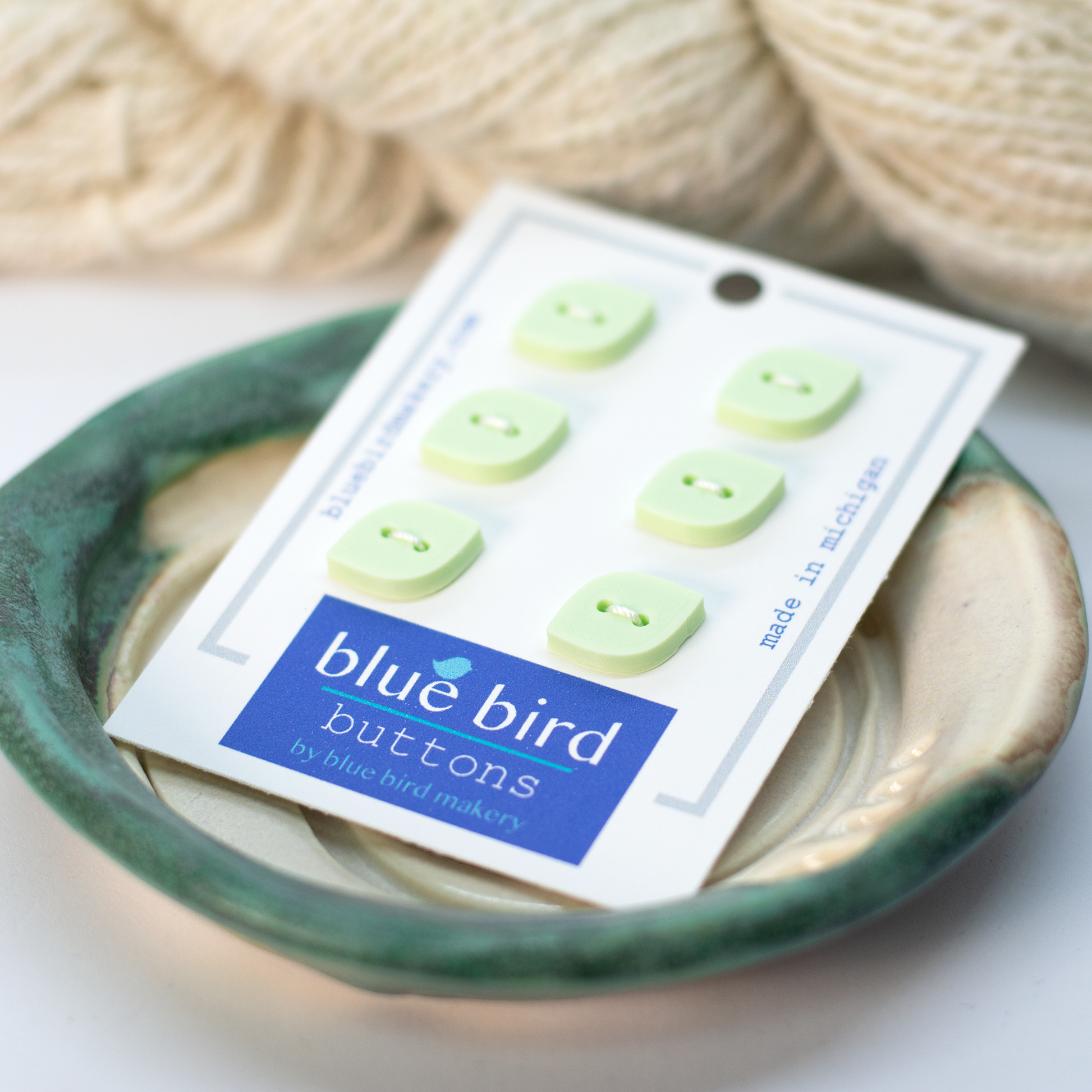 A pack of 12mm blue bird buttons in light green, displayed on a ceramic dish with yarn in the background