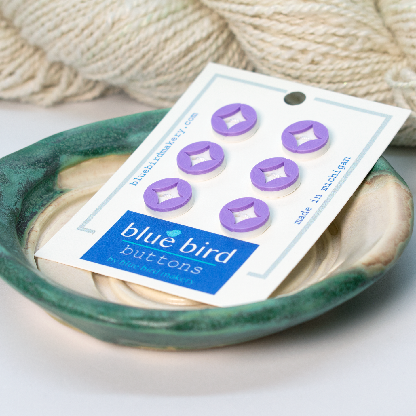 Blue Bird Makery craft supplies, featuring six purple buttons displayed on a ceramic dish