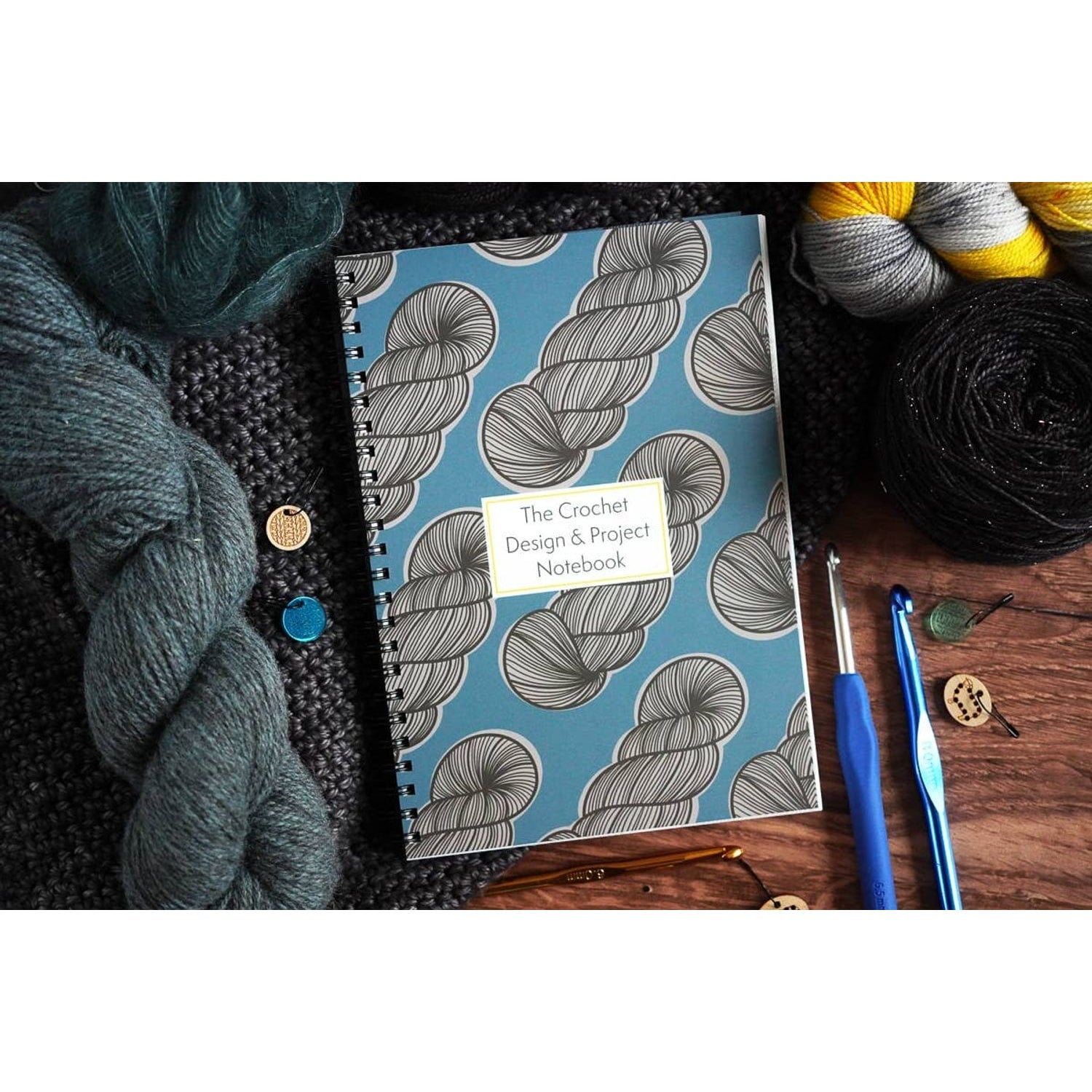 Spiral bound crochet pattern organizer notebook with blue yarn skein cover beside yarn balls crochet hooks and stitch markers