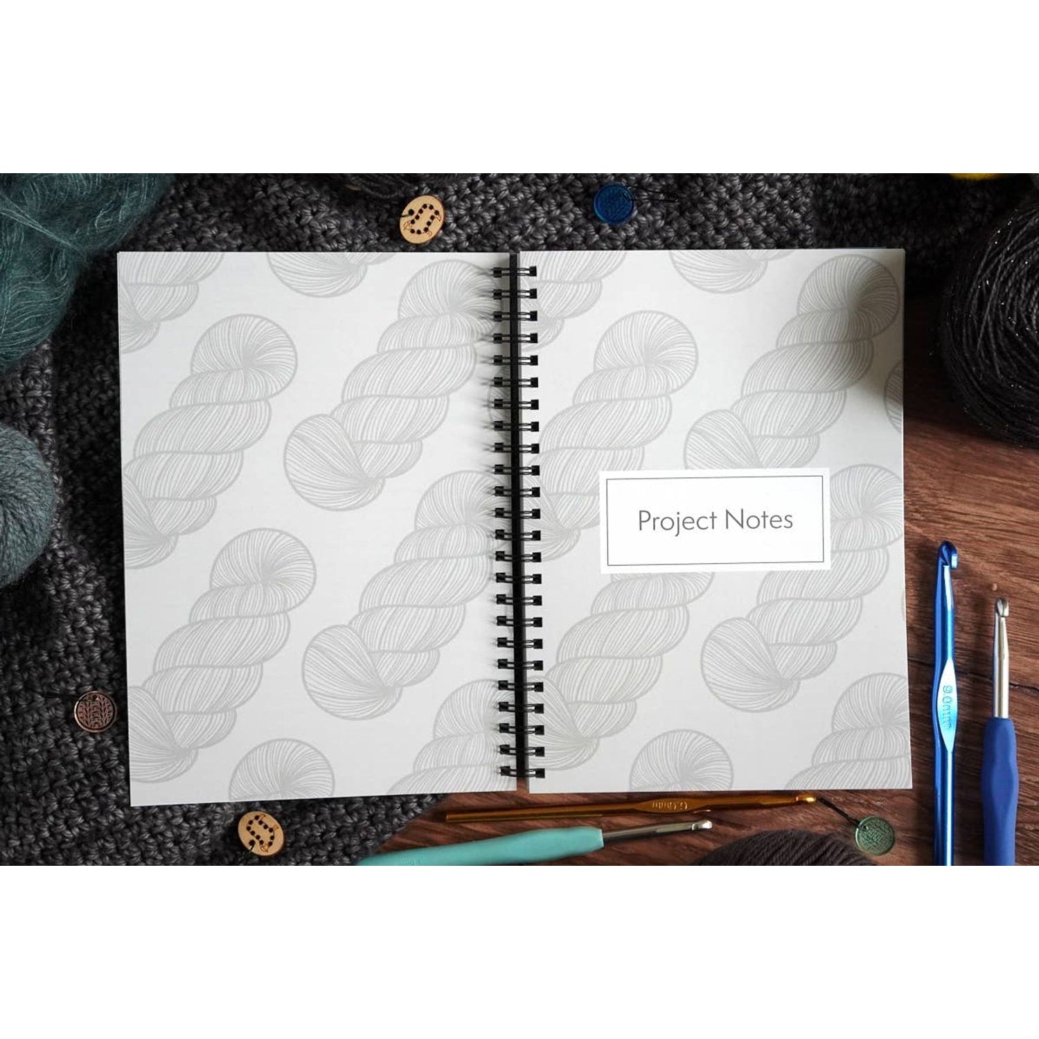 Open spiral notebook labeled Project Notes with yarn skein pattern beside hooks and yarn crochet pattern organizer notebook
