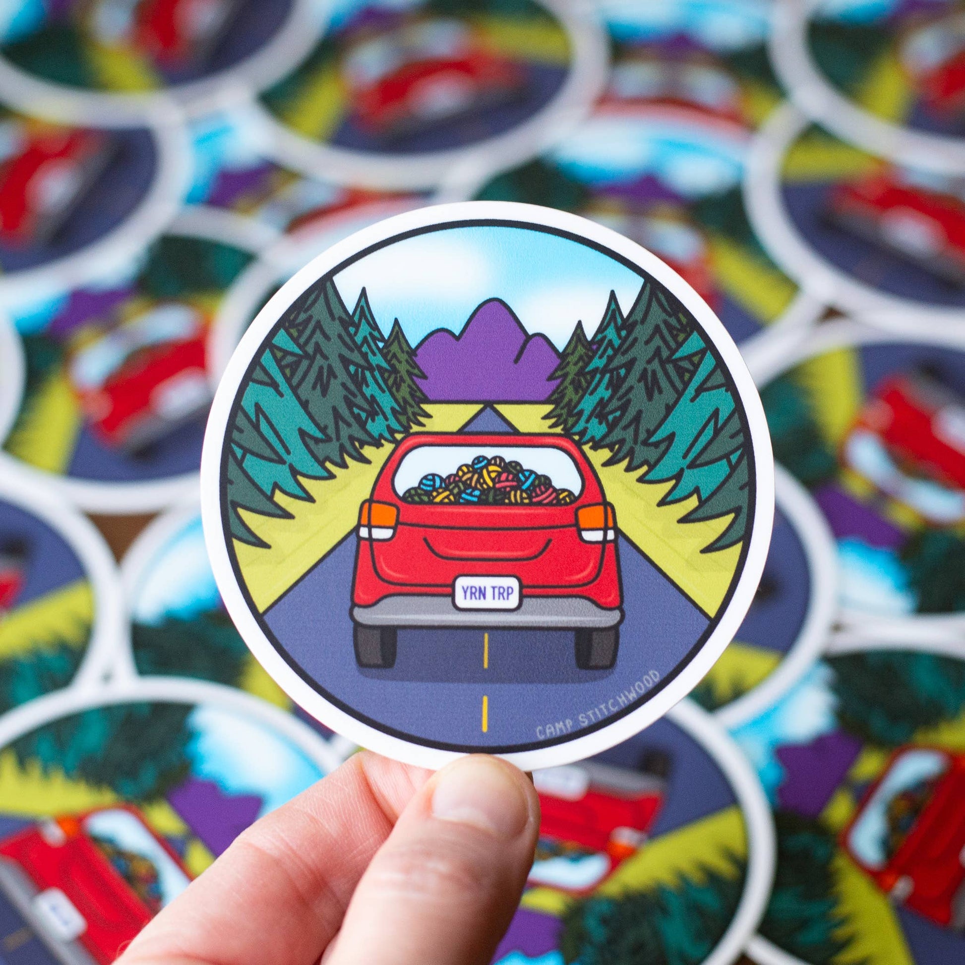 Camp sticker featuring a red car with yarn in the back on a scenic road surrounded by mountains