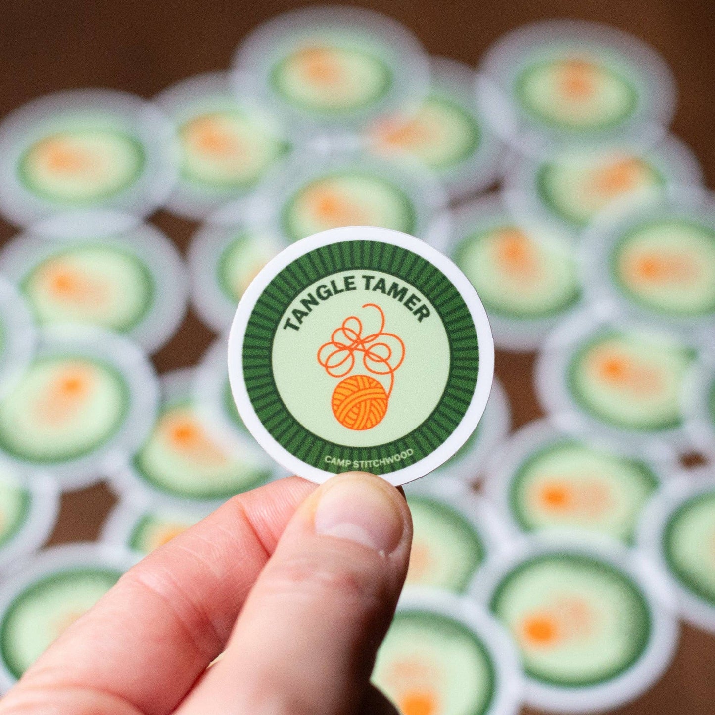 Hand holding a Camp Stitchwood name badge sticker featuring Tangle Tamer design with a green border