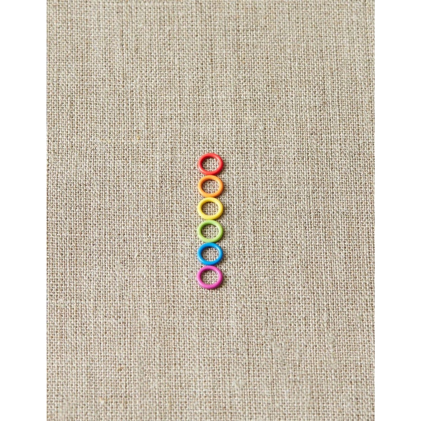 Cocoknits ring stitch markers in a rainbow of colors arranged vertically on a textured canvas background