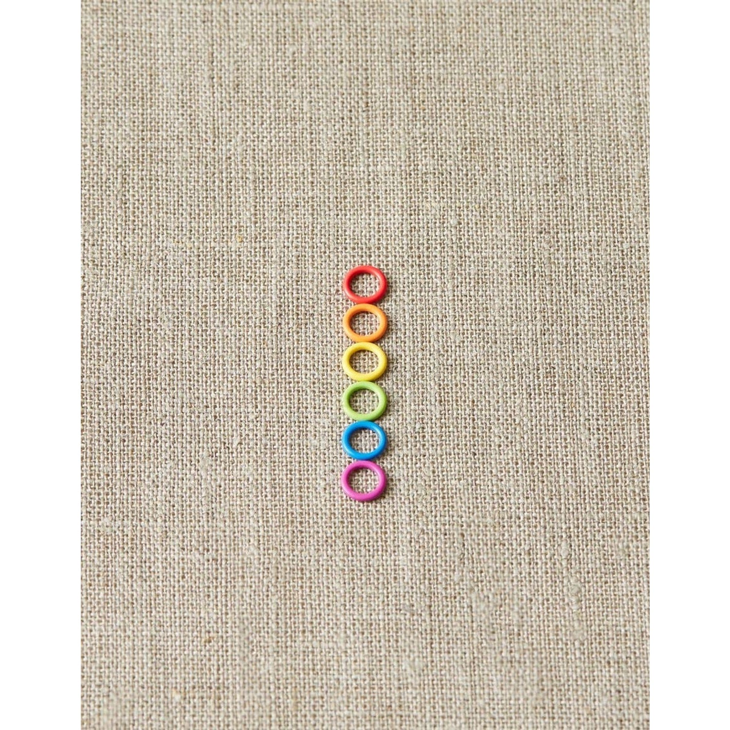 Cocoknits ring stitch markers in a rainbow of colors arranged vertically on a textured canvas background