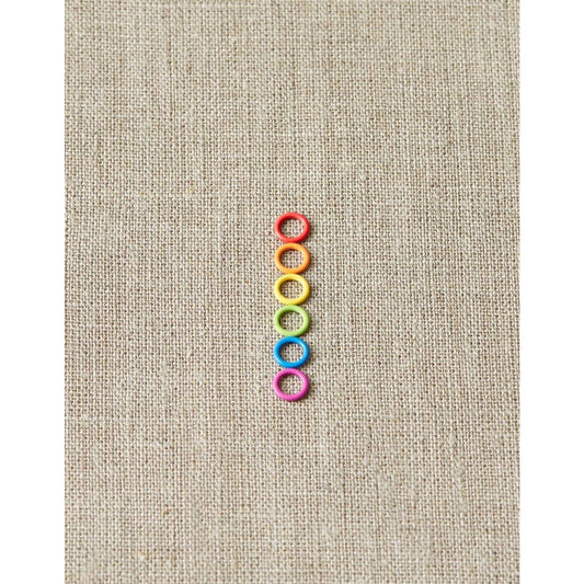 Cocoknits ring stitch markers in a rainbow of colors arranged vertically on a textured canvas background