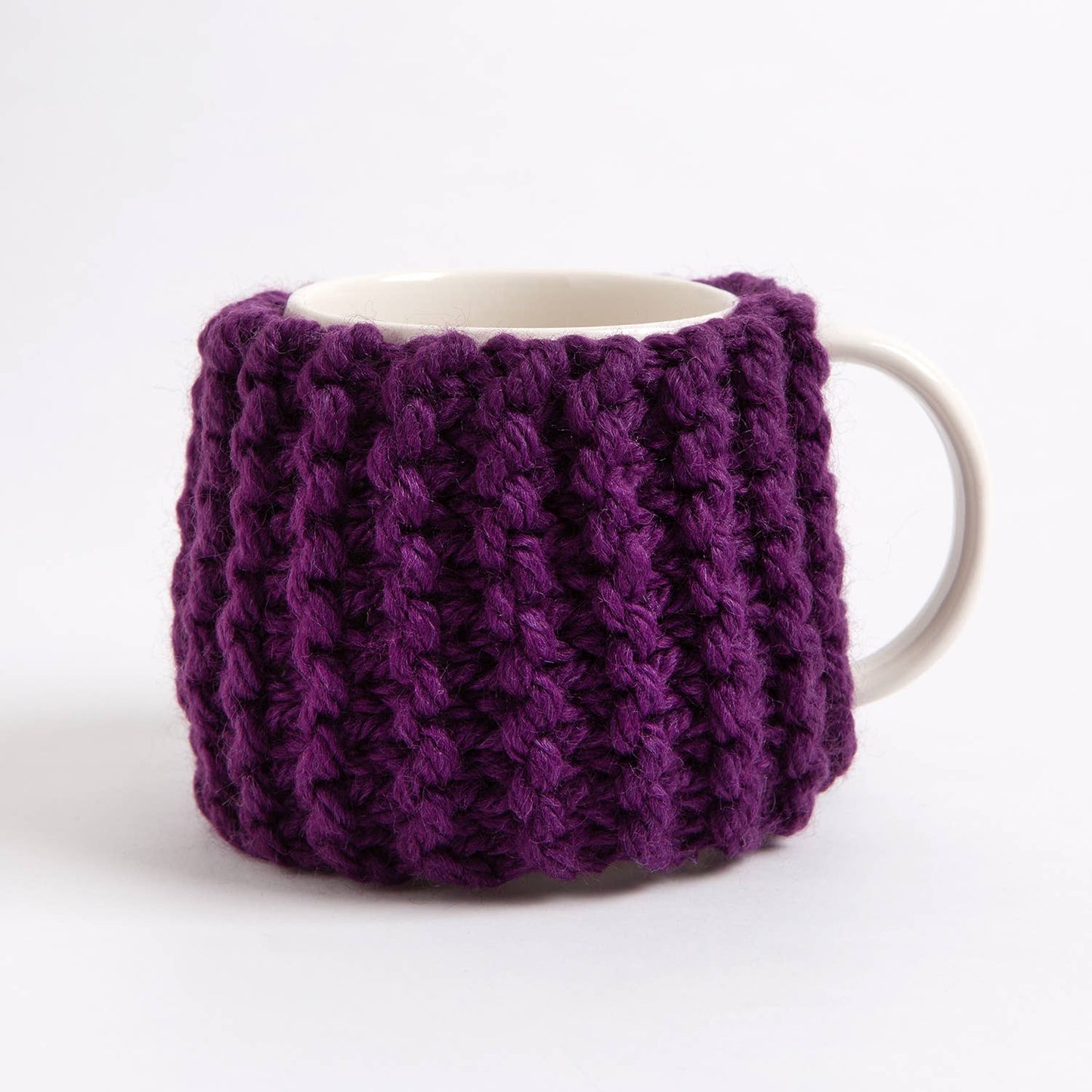 White ceramic mug wrapped in a chunky purple knit cozy shown as a beginner knitting kit for cup cozy