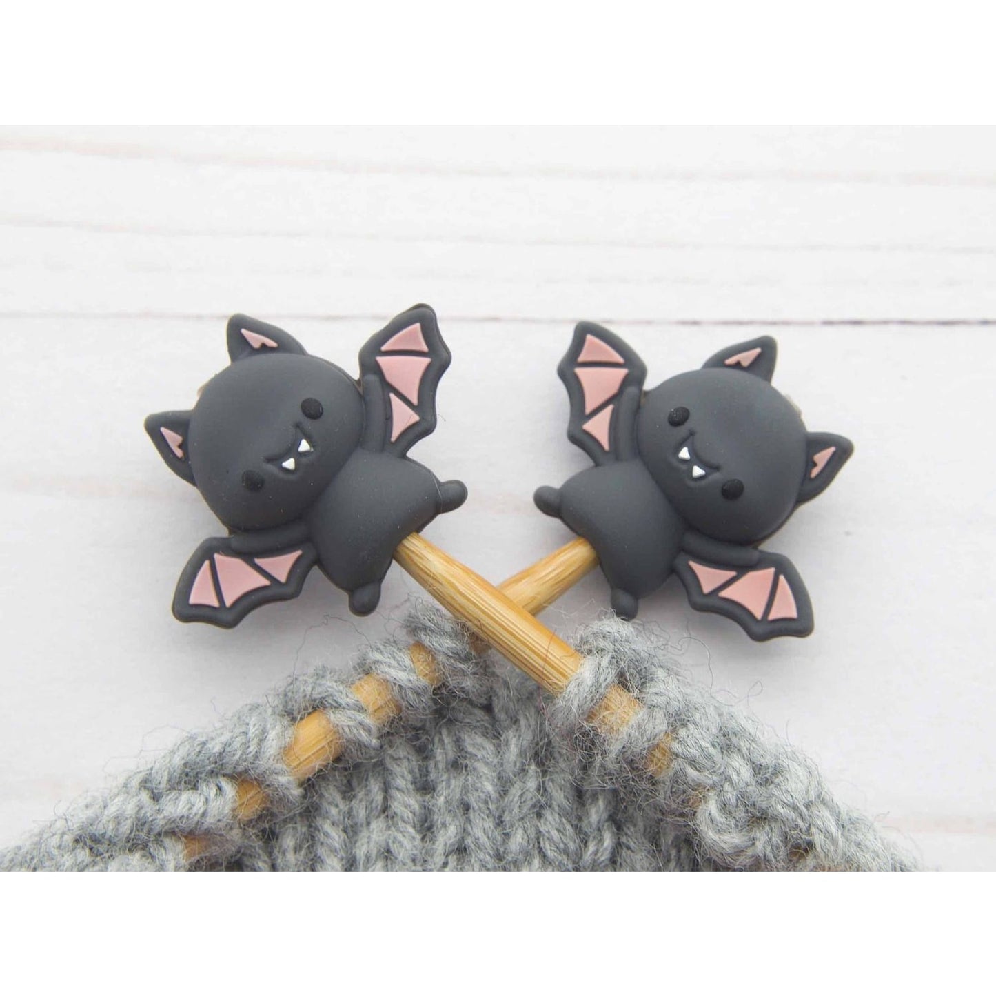 Two black bat stitch stoppers knitting on wooden needles securing live gray wool stitches in progress