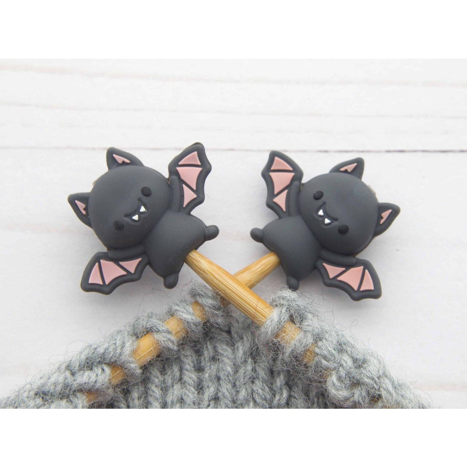 Two black bat stitch stoppers knitting on wooden needles securing live gray wool stitches in progress