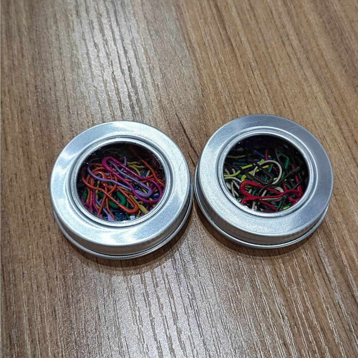 Two round metal tins filled with colorful stitch markers for crocheting from AriYARN Shop and Studio