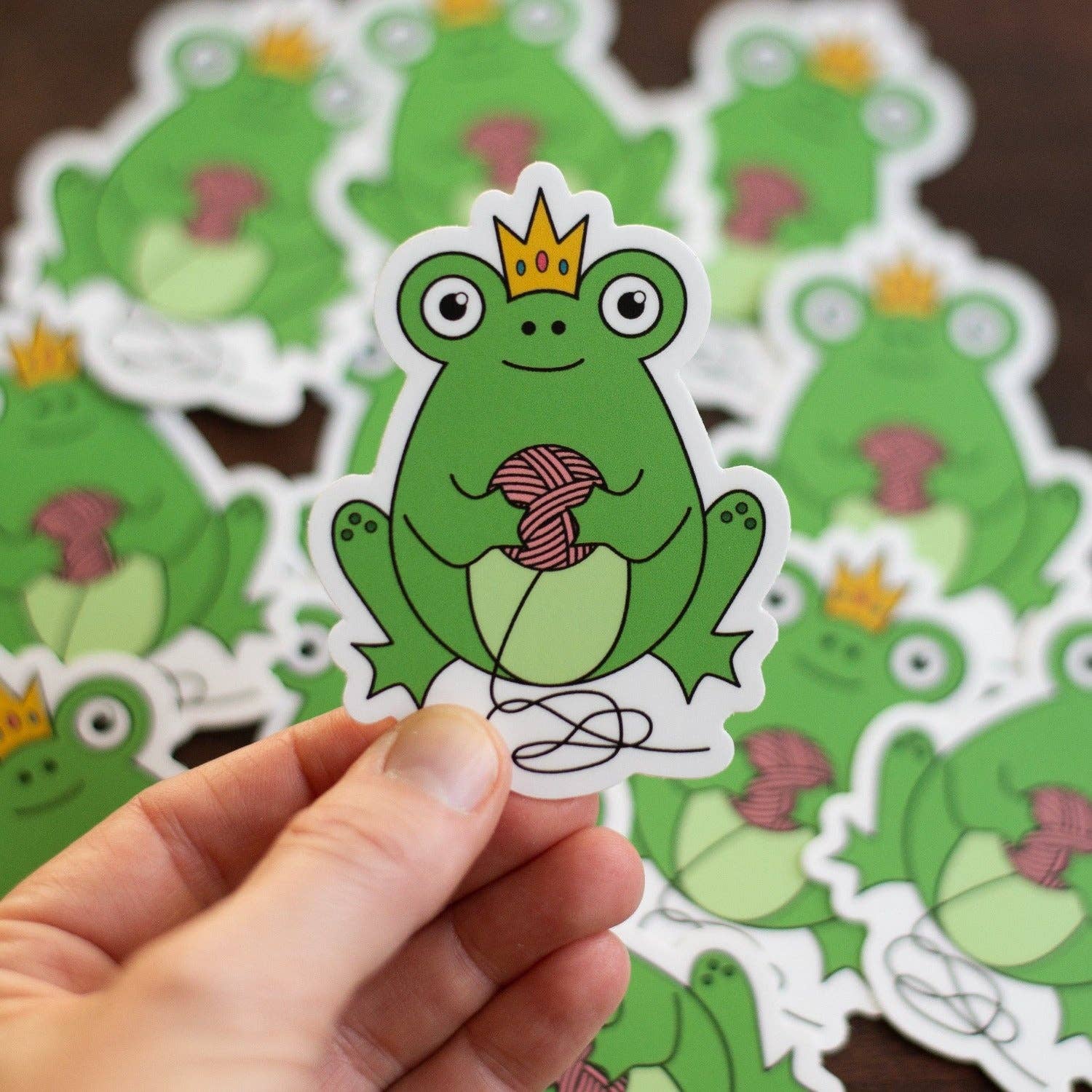Cute frog sticker with a crown, holding yarn, designed by Camp Stitchwood