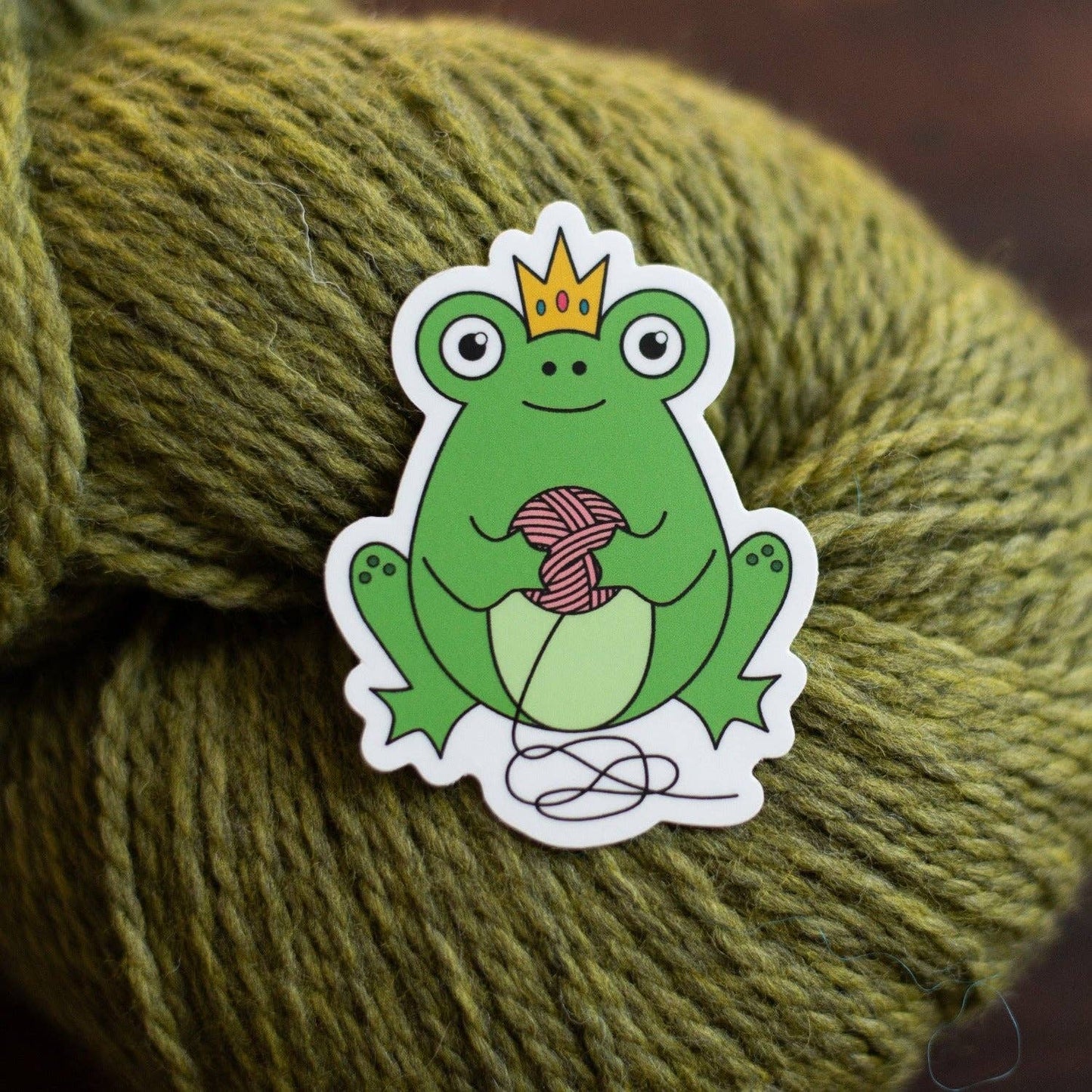 Cute frog sticker featuring a frog with a crown holding yarn, perfect for crafting enthusiasts