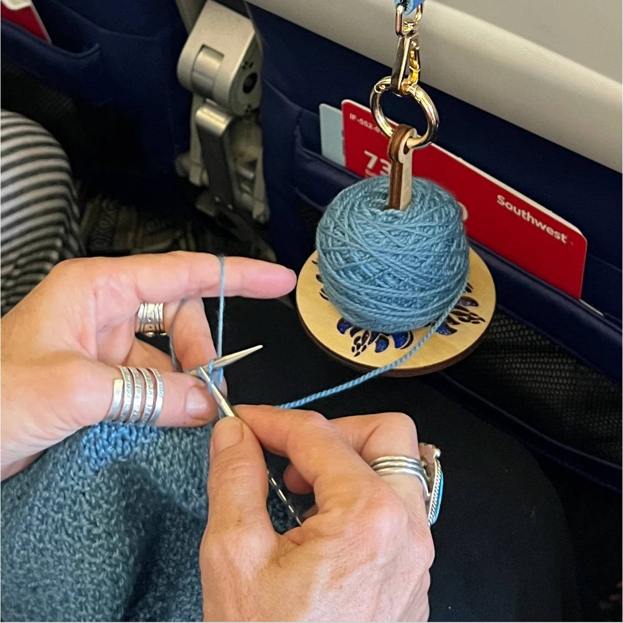Lemonwood wooden yarn holder keychain with blue yarn ball hanging on airplane seat while hands knit