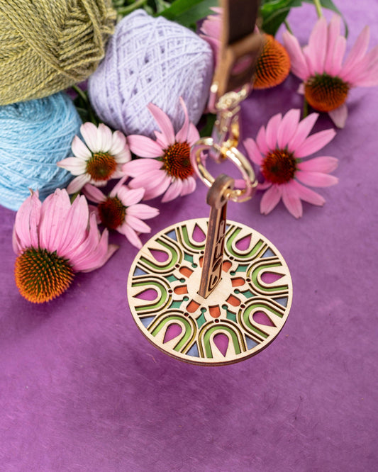 Lemonwood round wooden pendant keychain with colorful geometric inlay beside yarn and pink coneflowers