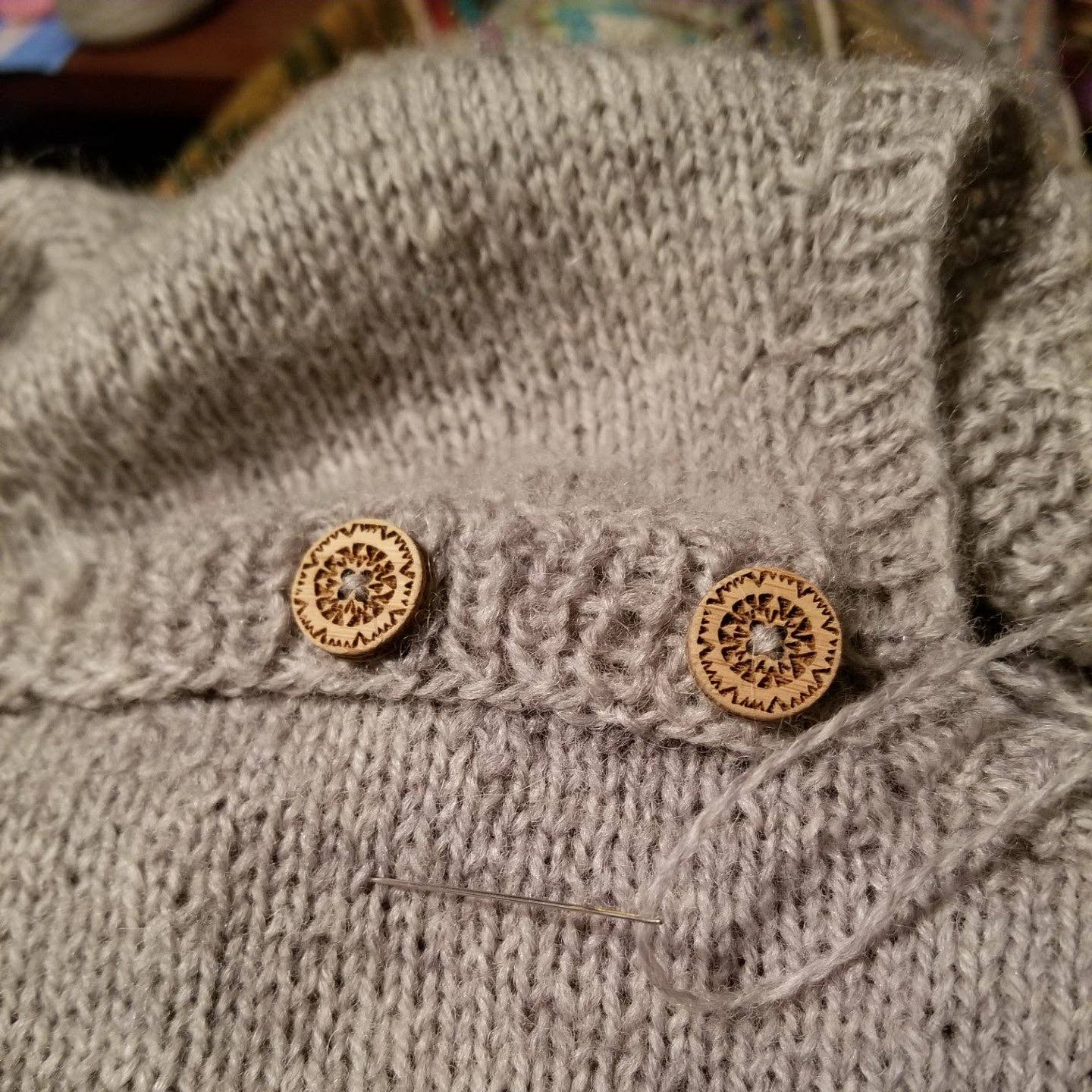 Katrinkles wooden buttons sewn on a light gray knitted sweater with a sewing needle nearby