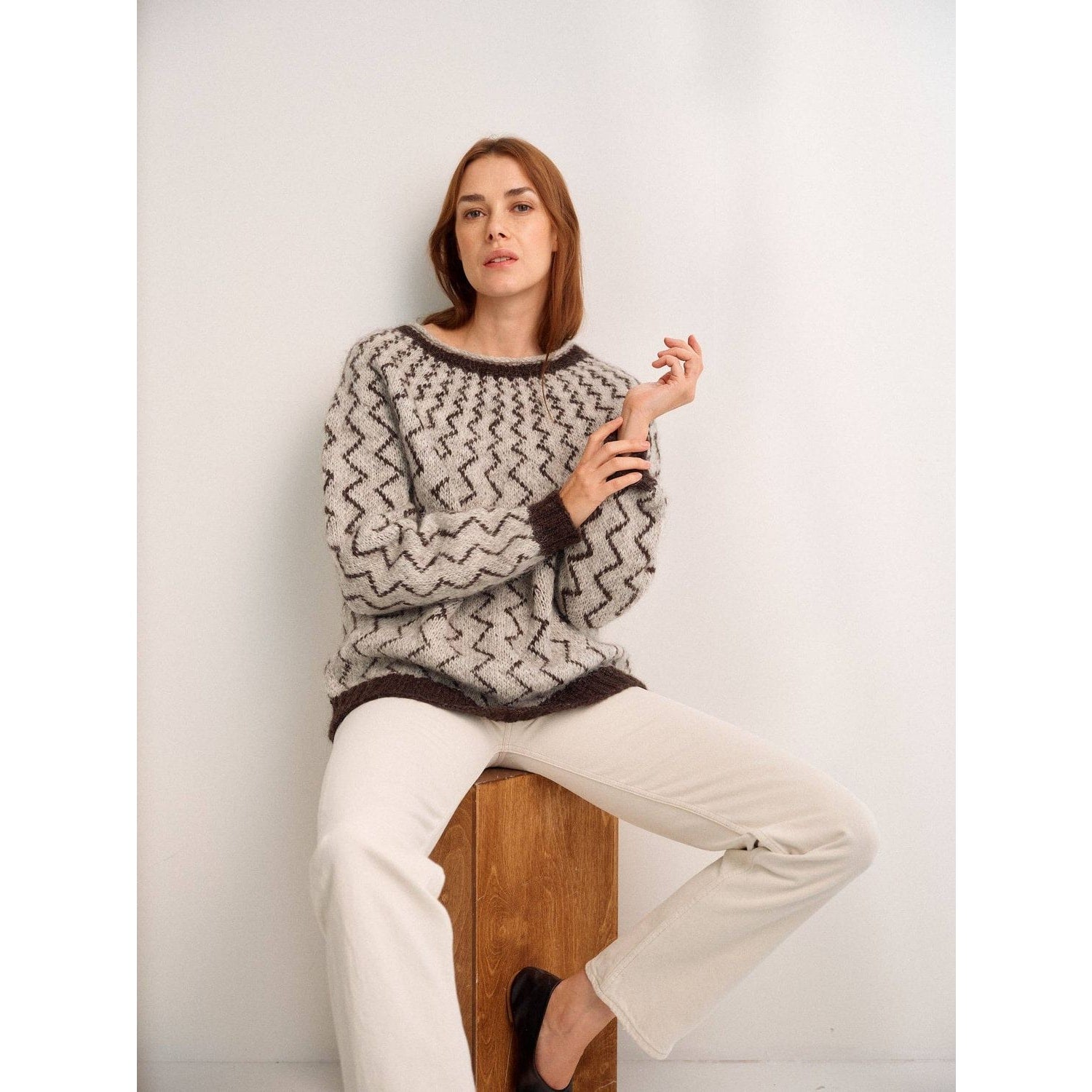 Stylish woman wearing a cozy knitted sweater showcasing unique patterns in knitting, seated on a wooden stool