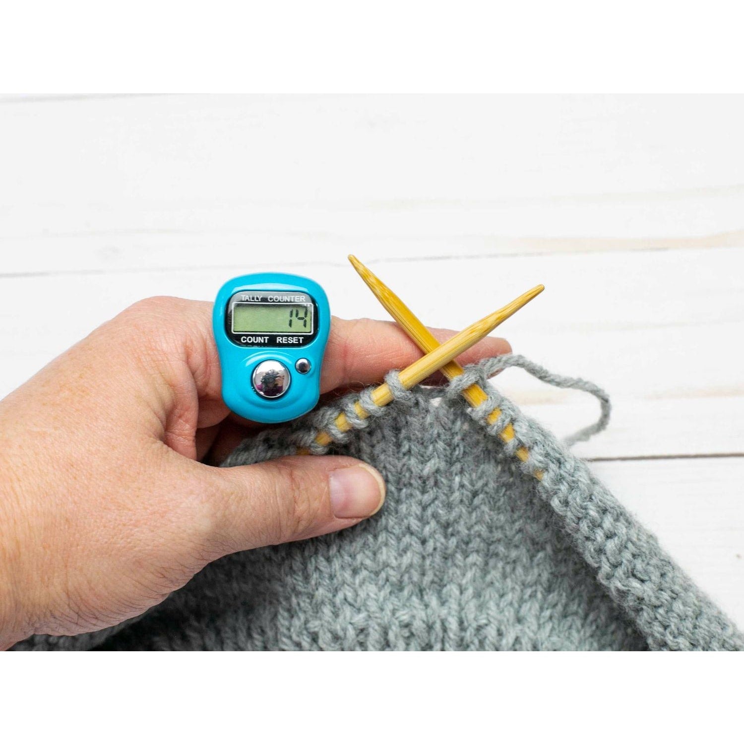 Hand holding a blue knitting row counter with wooden needles and gray yarn in the background