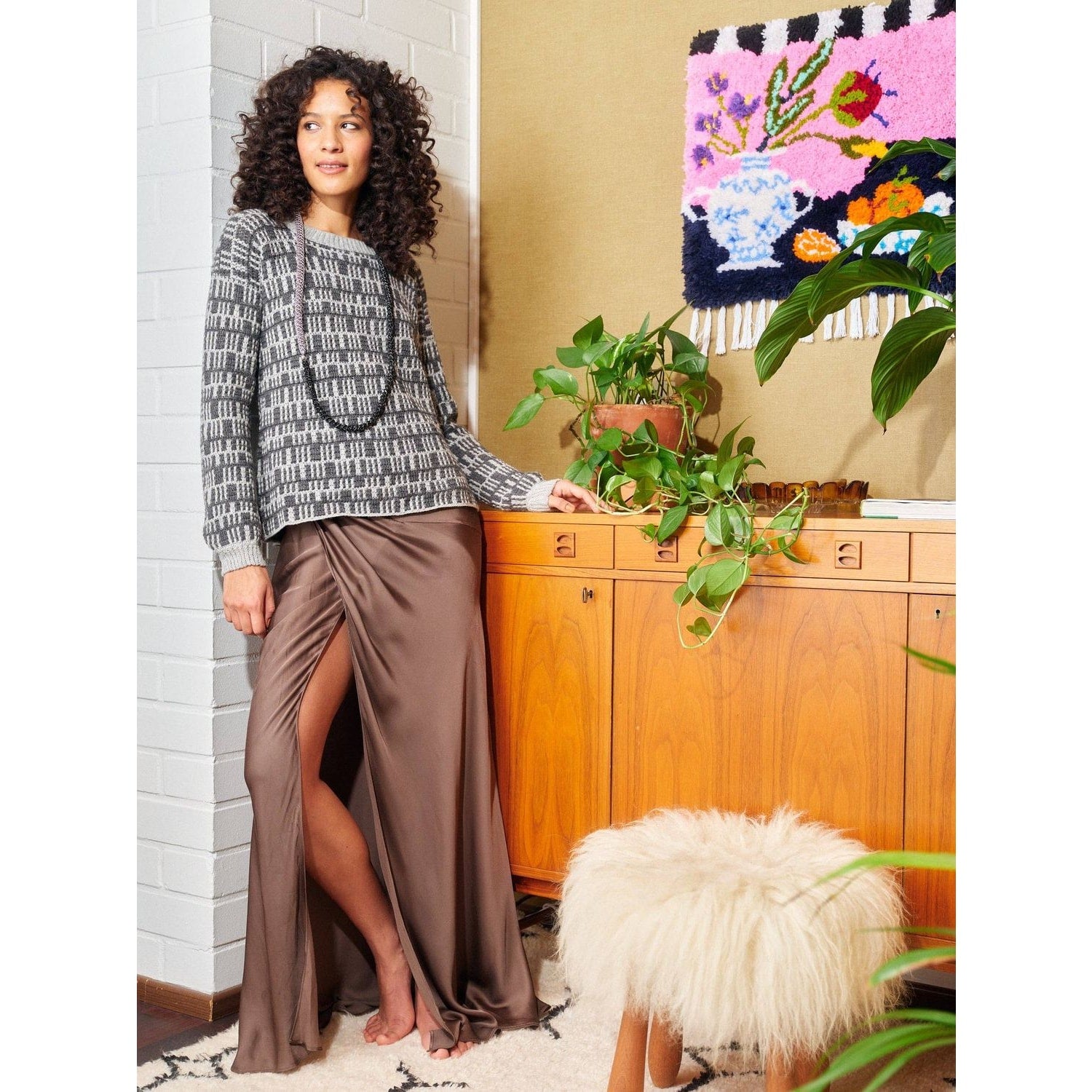 Woman wearing a stylish sweater and satin skirt, surrounded by plants and art, showcasing crochet with patterns