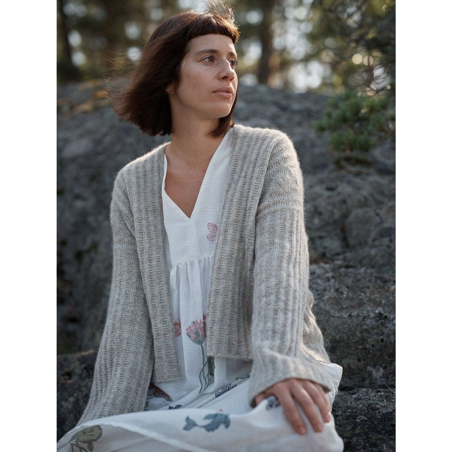 Woman wearing a cozy beige cardigan from Laine Magazine, seated on rocks in a serene outdoor setting