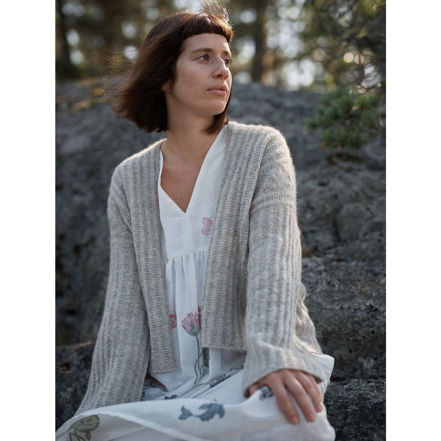 Woman wearing a cozy beige cardigan from Laine Magazine, seated on rocks in a serene outdoor setting