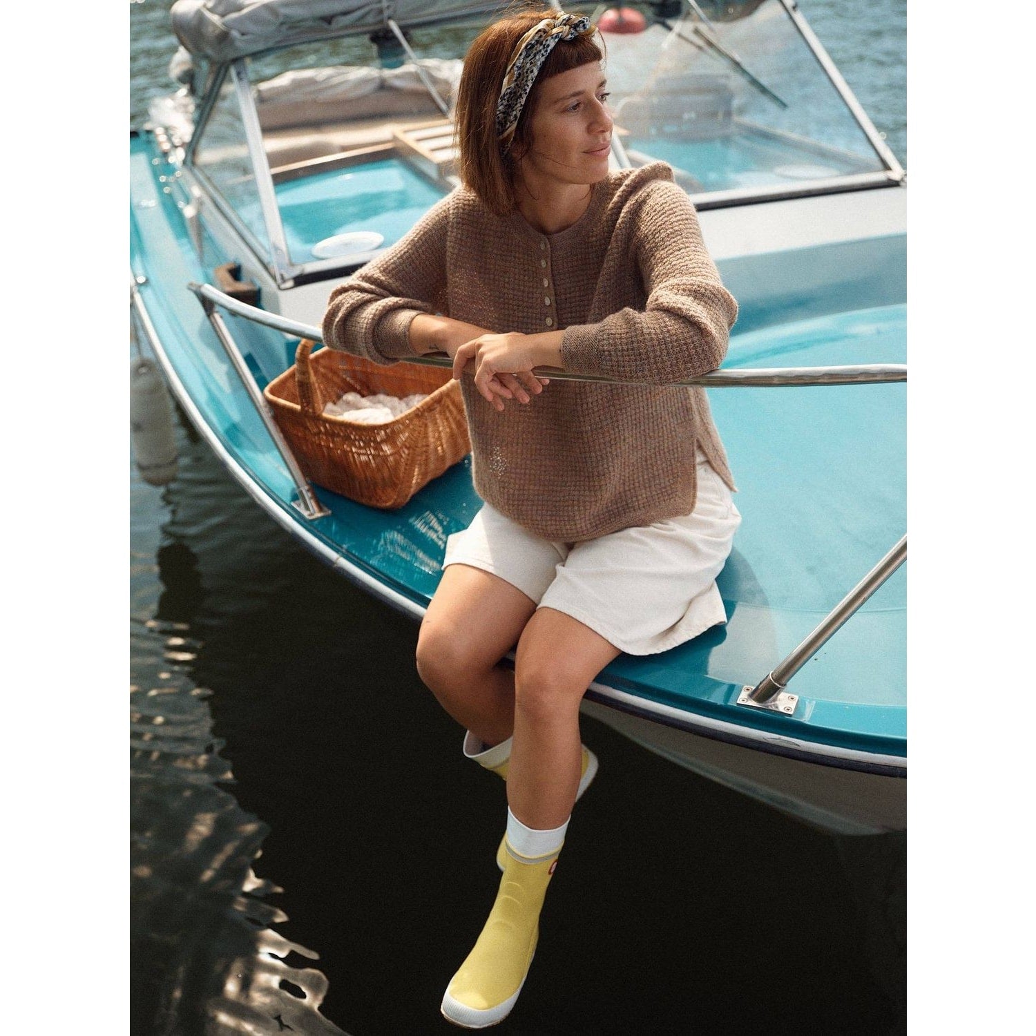 Woman in a beige sweater and yellow rain boots sitting on a boat holding Laine magazine
