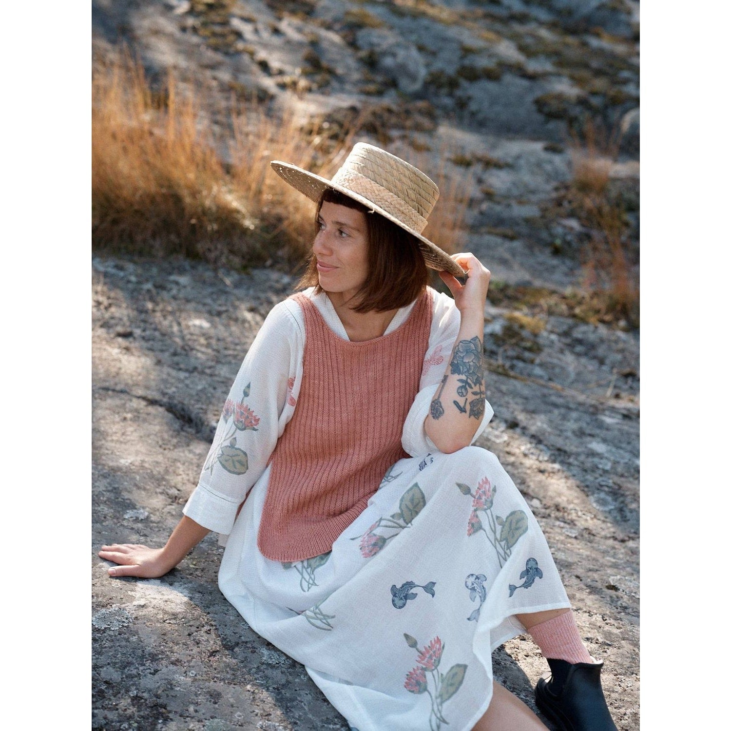 Woman in a straw hat wearing a Laine Magazine outfit, sitting on a rocky surface surrounded by nature