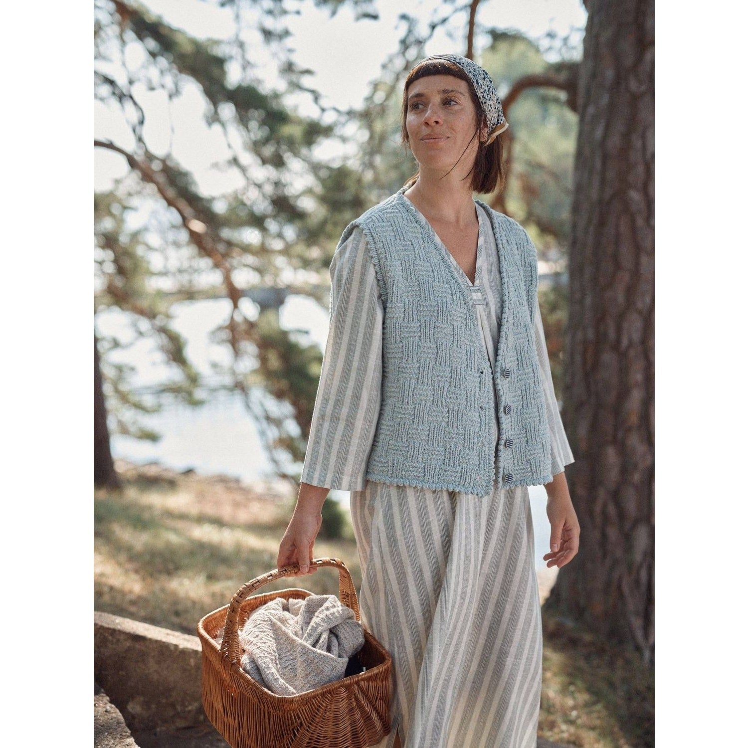 Woman wearing a striped dress and blue knitted vest from Laine Magazine, holding a basket outdoors
