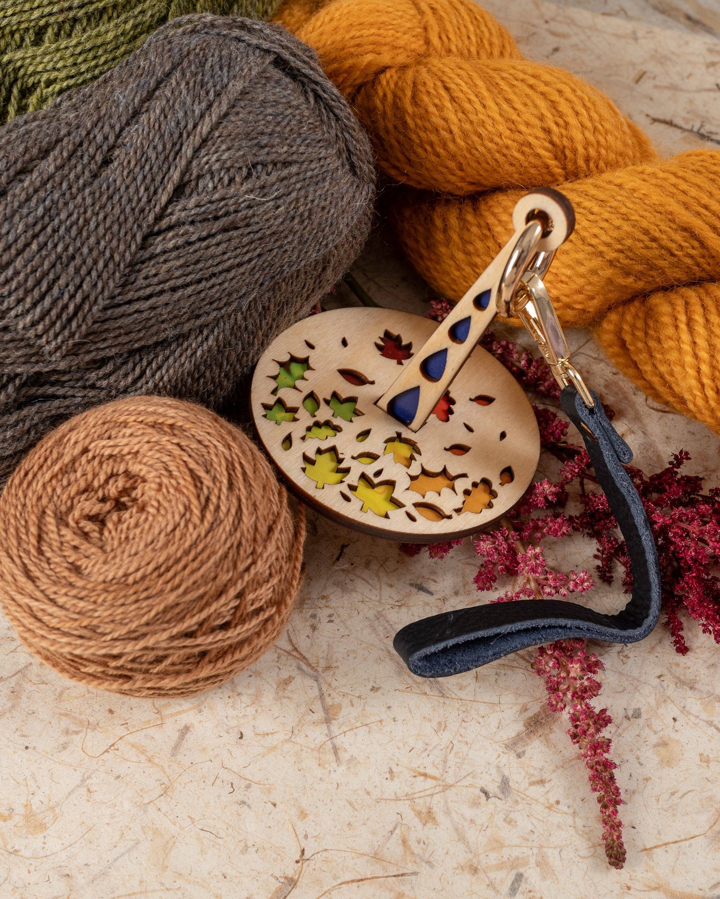 Lemonwood Autumn Tour stitch marker wooden key fob with leaf cutouts and leather strap beside mustard yellow gray and tan yarns