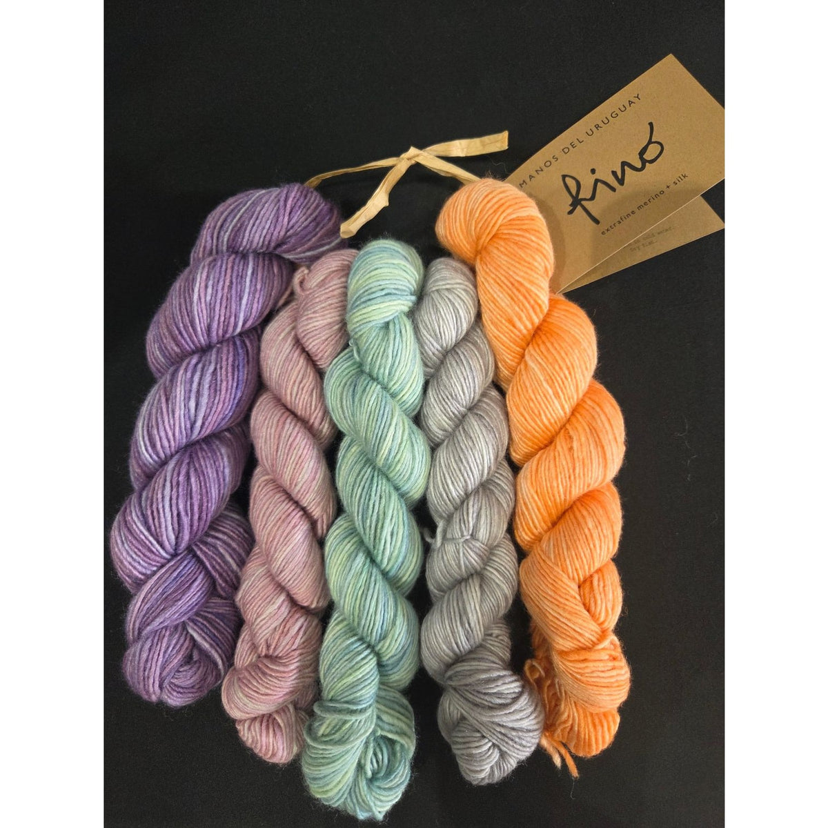 Five skeins of Manos del Uruguay Fino yarn in purple, teal, grey, and orange shades for knitting projects