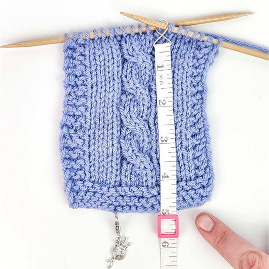 Blue knitted swatch with cable design and measuring tape showing precise measurements for knitting projects