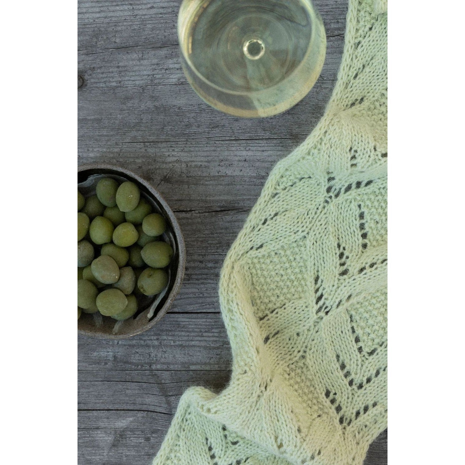 Light green knitted scarf pattern to knit displayed on a wooden table next to grapes and a glass of wine
