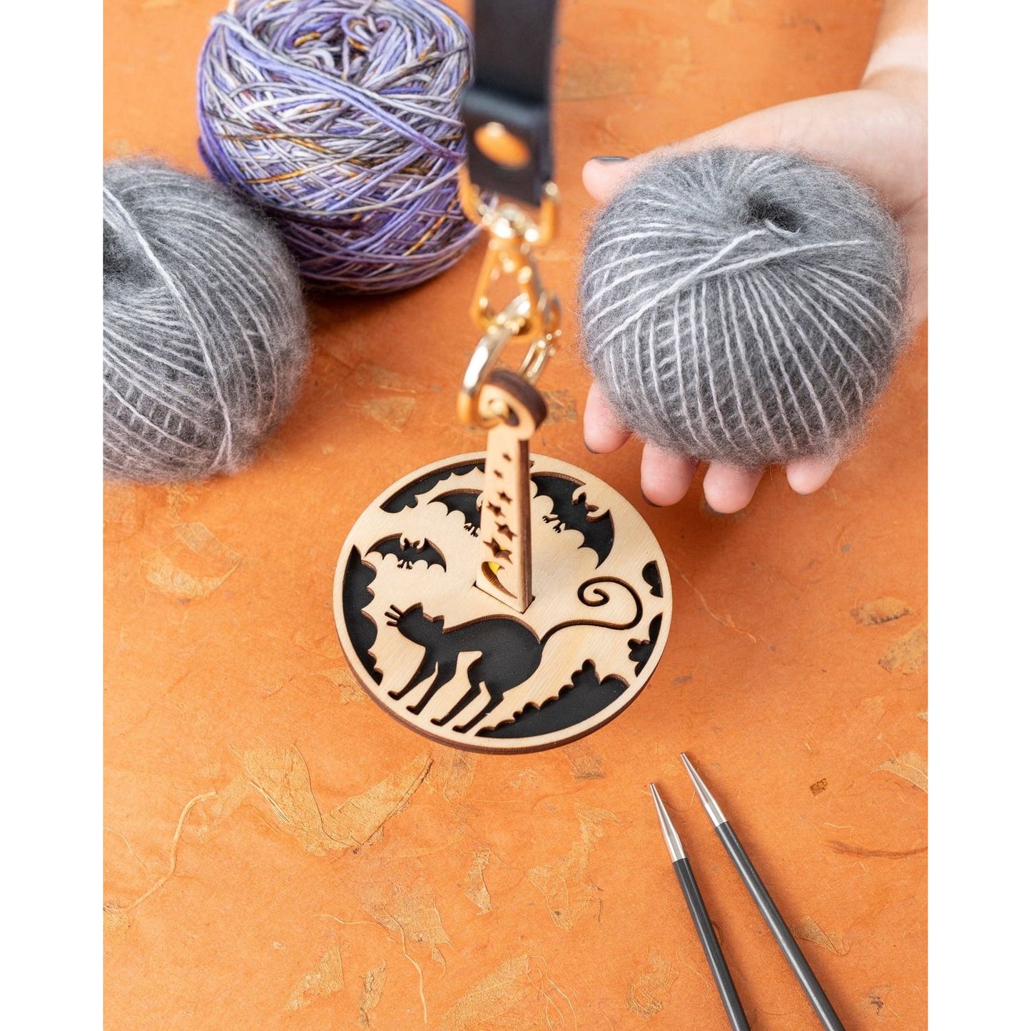 Lemonwood Something Familiar yarn with wooden cat bag tag and knitting needles beside gray and purple yarn balls