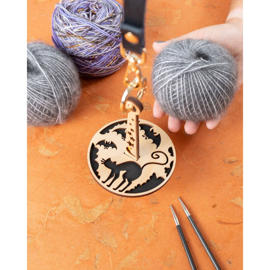 Lemonwood Something Familiar yarn with wooden cat bag tag and knitting needles beside gray and purple yarn balls