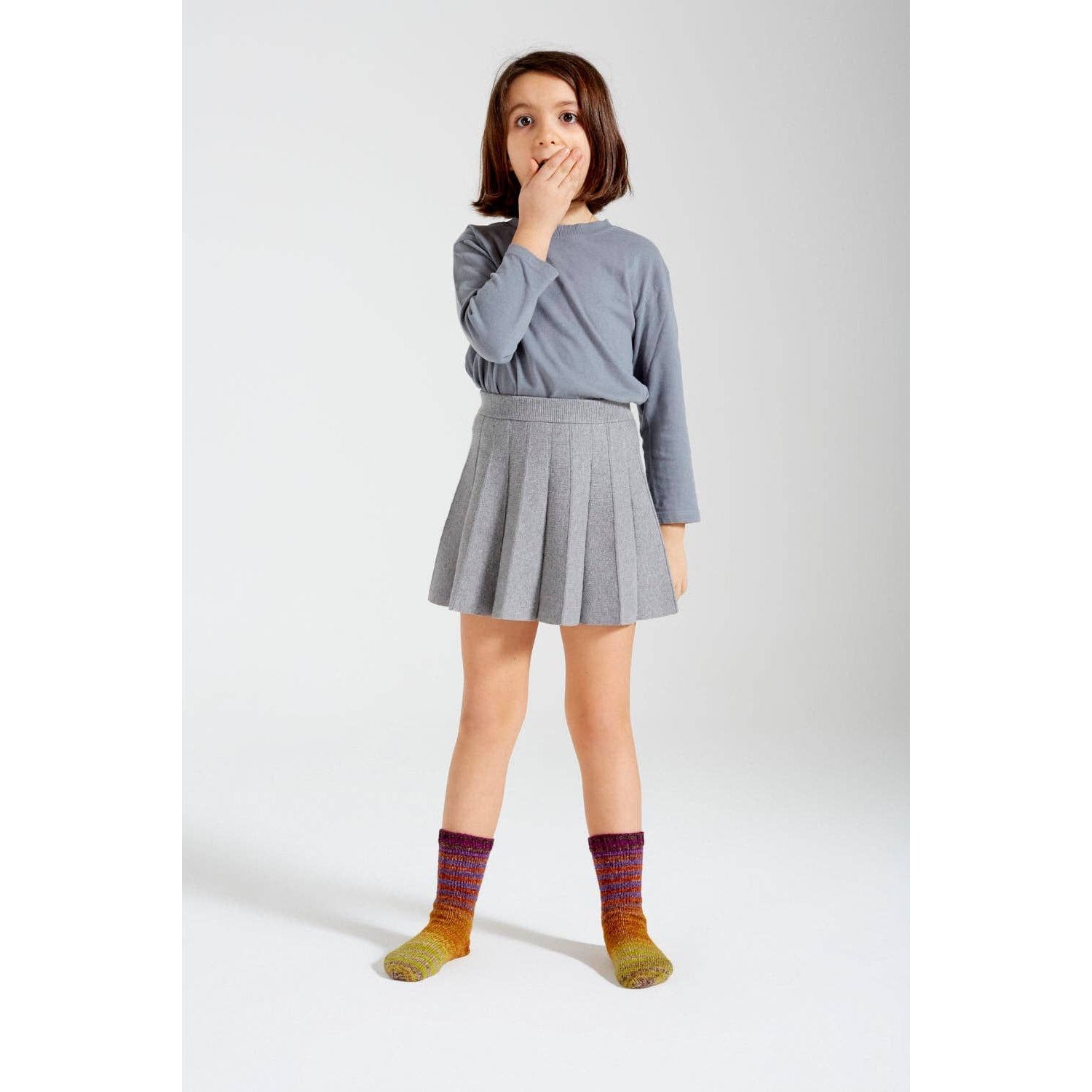 Child wearing knitted socks made from Urth Yarns Uneek Sock Kit Mini, styled with a gray top and skirt.