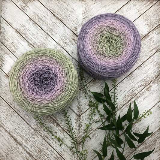 Two pastel green and lavender cake skeins of Wonderland Yarns Blossoms gradient yarn on whitewashed wood with greenery