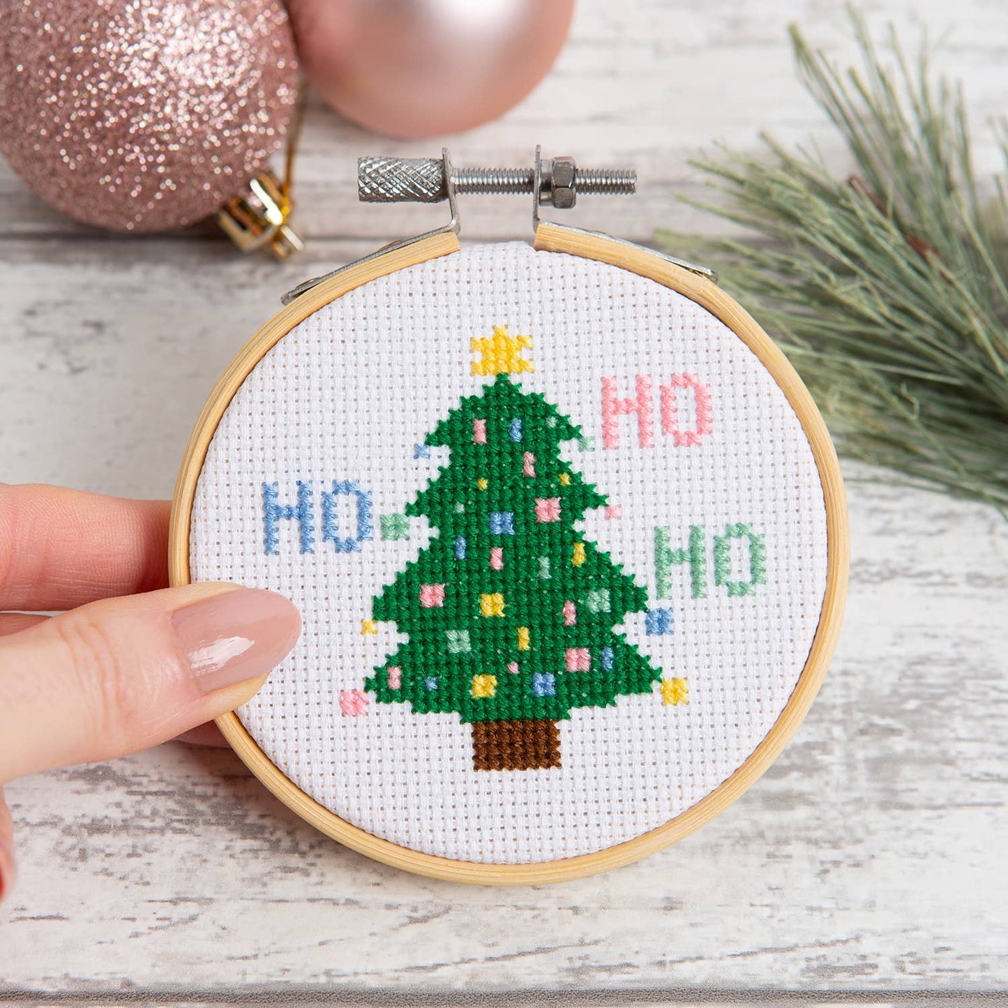 Hand holding small embroidery hoop with stitched Christmas tree and HO HO HO text Christmas tree cross stitch kit