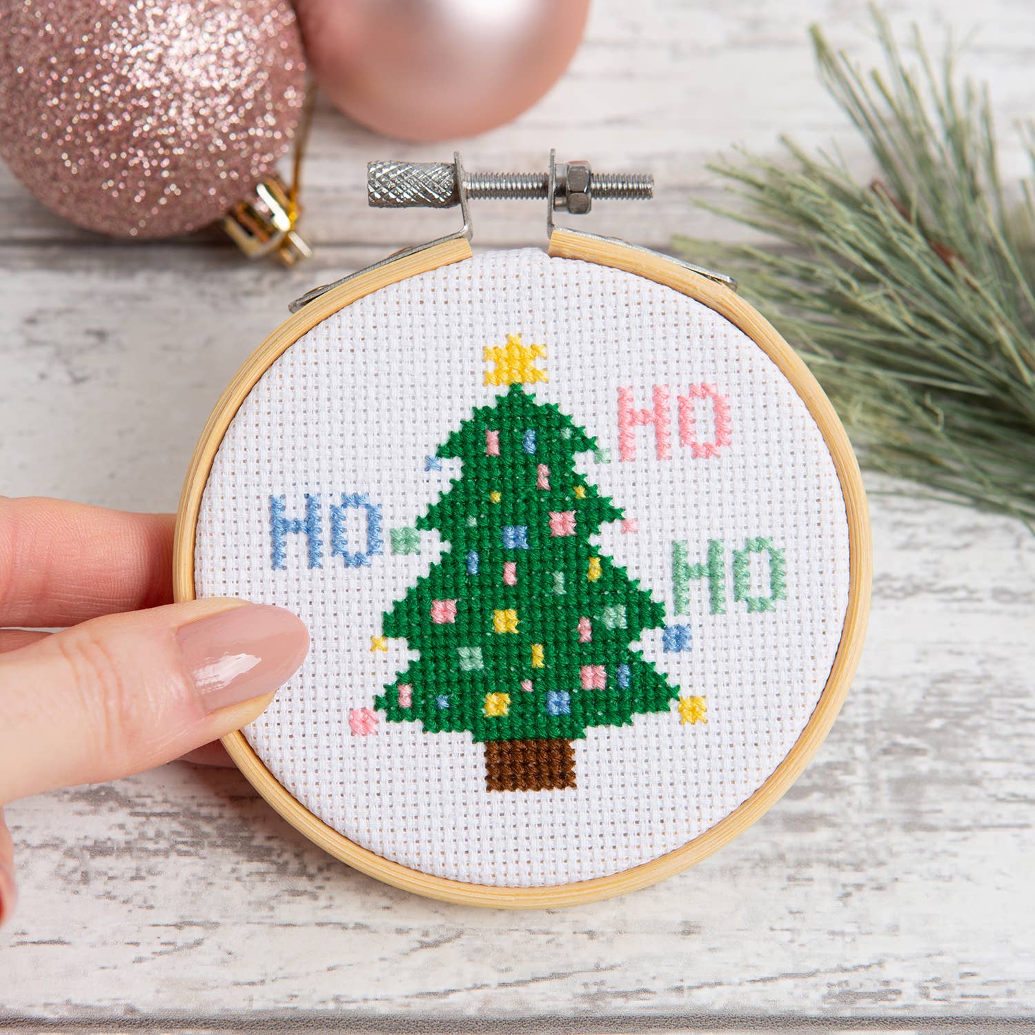 Hand holding small embroidery hoop with stitched Christmas tree and HO HO HO text Christmas tree cross stitch kit